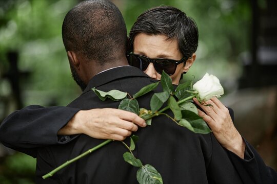 Medium shot of senior woman with white rose in hand giving comforting embrace to unrecognizable Black man grieving over loss of loved one at cemetery