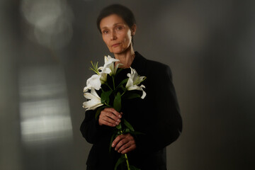 Waist up portrait of senior woman in black elegant suit holding white lilies grieving loss, while looking seriously at camera with stare expressing sadness on gray background in studio, copy space