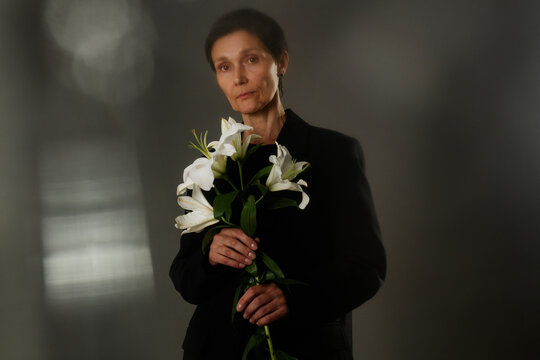 Waist up portrait of senior woman in black elegant suit holding white lilies grieving loss, while looking seriously at camera with stare expressing sadness on gray background in studio, copy space - Powered by Adobe