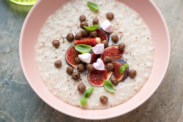 Oatmeal porridge served with fresh figs, chocolate balls and marshmallow in a roseate bowl, middle close-up, horizontal shot on a beige granite surface