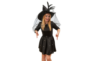 Smiling young woman in black witch hat and black dress points her fingers down. White background. Mockup. Halloween costume party