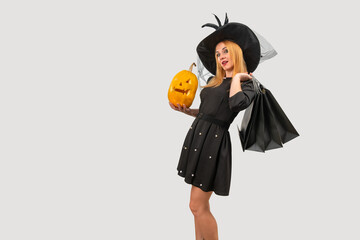 Blonde woman wearing a witch hat is holding a carved pumpkin and carrying shopping bags