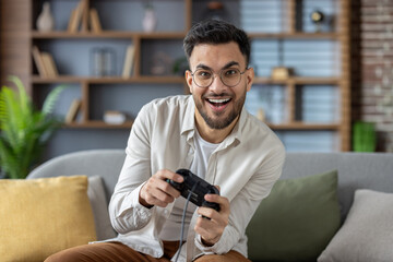 Portrait of an emotionally excited young Indian man sitting on the couch at home and playing online...