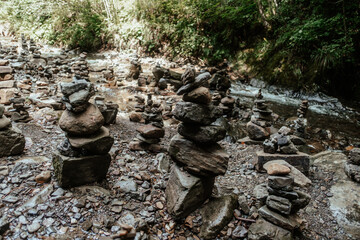 Visitors arrange various stones into towering cairns along a tranquil riverbank surrounded by lush greenery in a serene natural setting