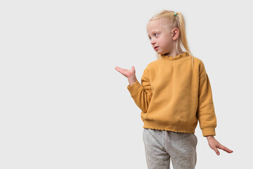 Adorable little girl with blond hair in yellow hoodie and gray pants showing something on empty copy space isolated on white background