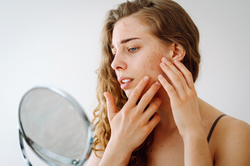 A young woman with curly hair is inspecting her face in a mirror, concentrating on blemishes and...