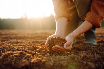 A young farmer kneels on the ground at sunset, cradling a handful of dark, fertile soil while surrounded by a landscape, highlighting the connection to nature. Gardening and ecology concept.