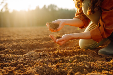 A young farmer kneels on the ground at sunset, cradling a handful of dark, fertile soil while...