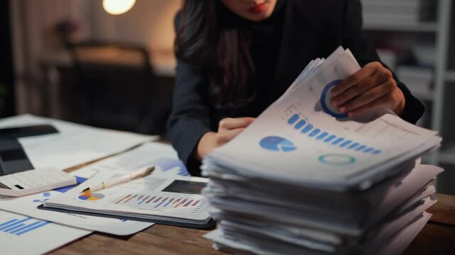 Businesswoman analyzing financial data and charts while working late at night, showcasing dedication and focus in a corporate environment filled with paperwork and deadlines