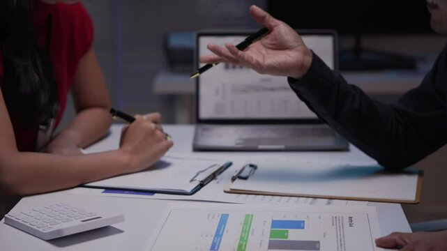 Business partners shaking hands after signing contract in office at night. They are working together using laptop computer and financial document