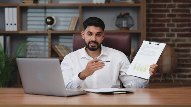 Serious indian office worker gesturing with hands while sitting at table with portable laptop. Earnest businessman pointing with pen on clickboard with paperwork.