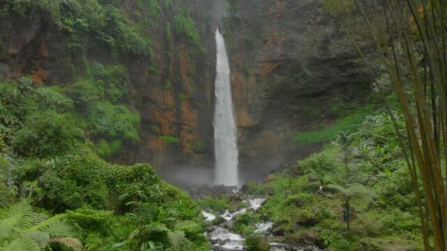 Aerial view of kapas biru waterfall cascading into a serene river surrounded by lush jungle and rocks, lumajang, indonesia.