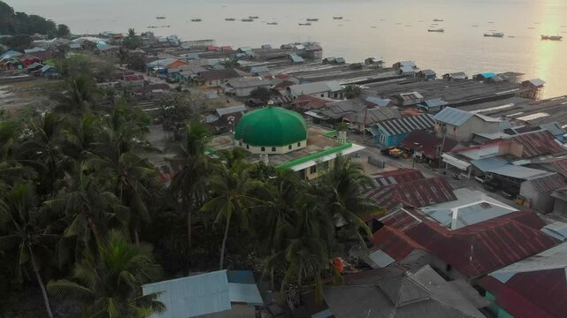 Aerial view of a tranquil coastal village with a green dome mosque and fishing boats at sunset, Tanjung Binga, Indonesia.