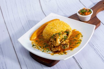 A vibrant, traditional Ecuadorian dish, Seco de Pollo, served with flavorful yellow rice and fried plantains. The dish features a tender chicken leg simmered in a rich, aromatic sauce made from tomato