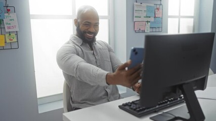 Young man taking a selfie in an office, smiling at his phone while seated at a desk with a computer and motivational notes on the wall.