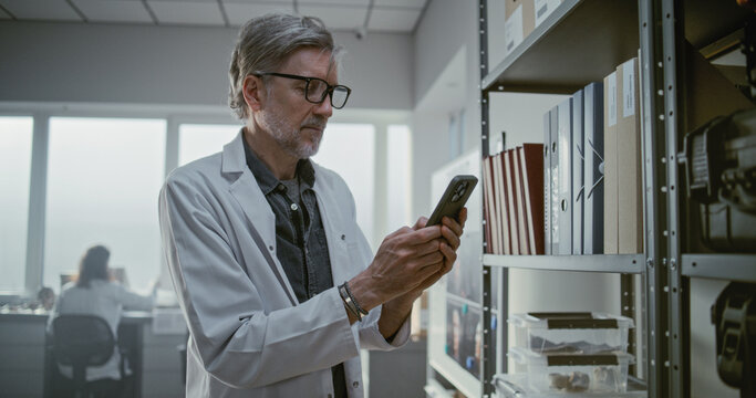 Archaeological laboratory: Mature scientist chats or surfs the Internet using mobile phone while standing near shelf with folders and boxes with fossils. Female colleague works in the background.
