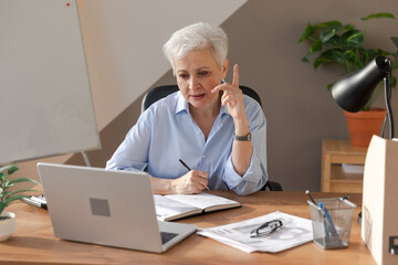 Middle aged senior woman using laptop computer writing notes at workplace. Focused mature old businesswoman work at office making written records doing online work. Mature boss professional worker