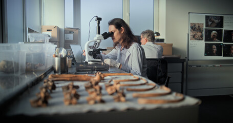 Female scientist analyzes sample of fossil remains under microscope. Bones of prehistoric human skeleton lying on table. Microbiologists or archaeologists work in advanced archaeological laboratory.