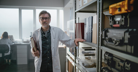 Modern archaeological science laboratory: Portrait of mature scientist looking at camera and holding folder while standing near shelf with boxes with fossils. Female colleague works in the background.