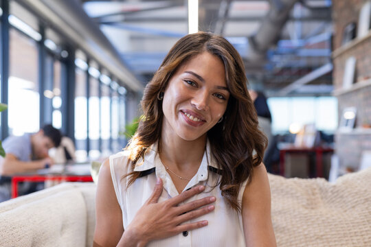 On video call, Smiling woman sitting on couch in modern office, hand on chest
