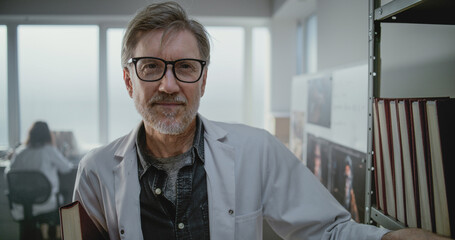 Modern archaeological science lab: Portrait of mature scientist in glasses and lab coat smiling and looking at camera while standing with folder near shelf. Female colleague works in the background.