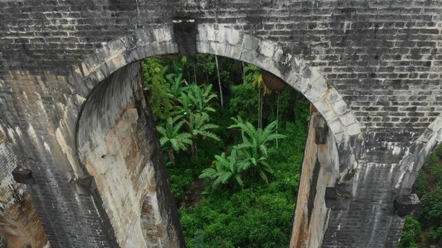 Aerial view of the nine arches bridge surrounded by lush tea fields and tropical plants under a cloudy sky, Ella, Sri Lanka.