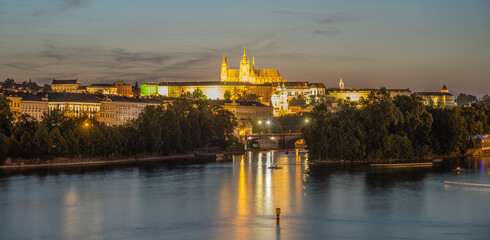 A nighttime view of Prague Castle illuminated across the Vltava River. The castle spires and buildings are reflected in the water, creating a picturesque scene. The cityscape is bathed in warm, golden