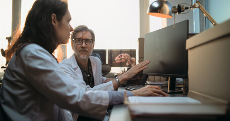 Two diverse scientists discuss 3D visualization of ancient human head anatomy displayed on PC and tablet computer screen, conduct historical research. Archaeologists work in archaeological laboratory.