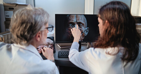 Two diverse scientists discuss 3D model of human head anatomy displayed on PC and tablet computer screens, conduct historical research. Archaeologists or paleontologists work in archaeological lab.