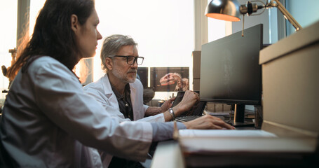 Two diverse scientists discuss 3D visualization of ancient human head anatomy displayed on PC and tablet computer screen, conduct historical research. Archaeologists work in archaeological laboratory.