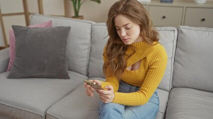 A young attractive woman with blonde hair in a yellow sweater looks concerned while holding her phone, sitting in her living room at home.