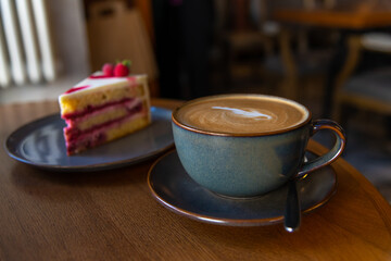 Side view of blue porcelain cup of cappuccino with latte art standing on brown wooden table in cafe or restaurant. Blurred raspberry cake in the background. Soft focus. Copy space. Food business theme