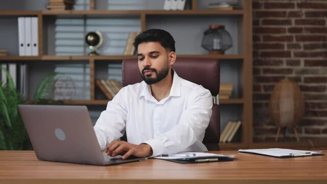 Front view of pleasant hindu male open portable laptop and starting doing work indoors. Smiling man dressed in formal attire sitting at personal table on brown leather chair.