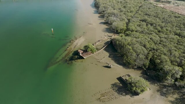 Aerial view of a serene ship graveyard with rusting shipwrecks along the coastline surrounded by lush green forest, Port Adelaide, Australia.