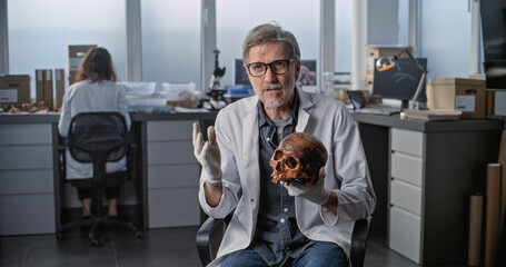 Mature scientist speaks to camera, shows fossil skull of ancient human during interview for popular...