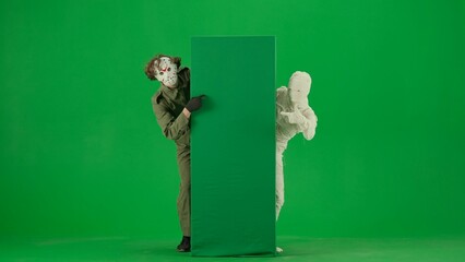 Portrait of person in a mummy bandage on chroma key green screen background. Mummy and maniac standing at empty board, pointing thumbs up.
