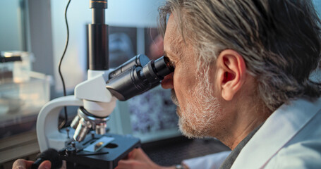 Mature microbiologist conducts fossil analysis under microscope, looks at microorganisms on laptop screen. Scientist works with advanced equipment in archaeological science laboratory. High Angle.