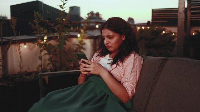 Portrait of crying young indian woman with smartphone sitting on the couch under the blanket and reading bad news email break up message from ex-boyfriend or personal problems at home terrace late