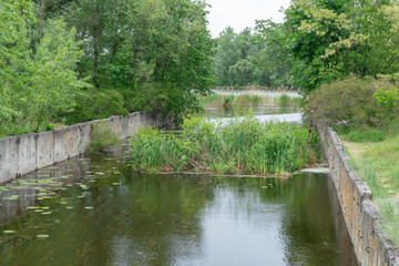 Support slabs along picturesque narrow river canal. Iron concrete reinforced pier from one bank of water to other. Trees bushes and reed grow along empty pedestrian. Reflection nature in river.