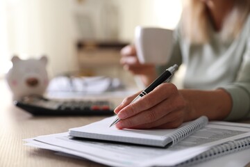 Fototapeta premium Budget planning. Woman with cup of drink taking notes at table indoors, closeup