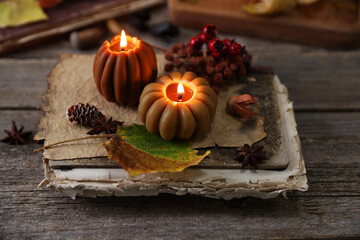 Burning candles in shape of pumpkins, old book and autumn decor on wooden table