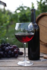 Red wine in glass, bottle, barrel and grapes on wooden table outdoors