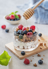 Natural greek yogurt with granola, honey, raspberries and blueberries in a glass on a light background
