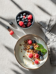 Natural yogurt with granola or muesli, raspberries and blueberries in a bowl on a light texture background with fresh berries and morning sunlight.