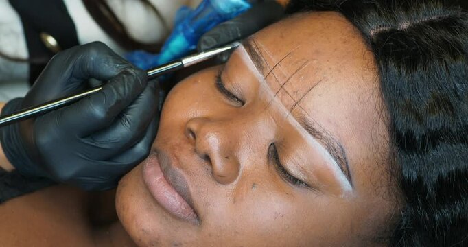 Beautician drawing white correction lines for eyebrow permanent tattoo on young black woman