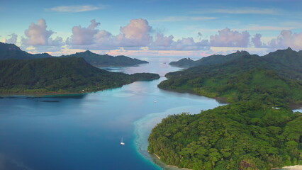 Tourist yacht in tropical ocean bay surrounded by lush green islands in French Polynesia, beautiful sunset sky in background. Remote wild nature paradise, exotic summer luxury travel. Aerial view