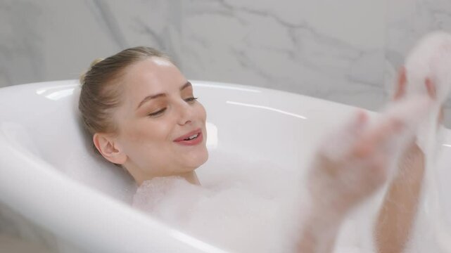 A young woman is enjoying a relaxing bath in a white tub filled with bubbles. She playfully blows the bubbles from her hand, creating a lighthearted and carefree moment.