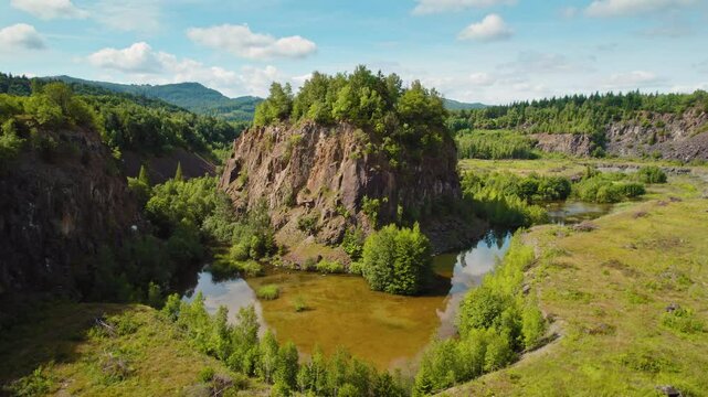Lush green hill rises from a valley, surrounded by a reflective pond and forested landscape under a clear blue sky. The Diabase quarry in Wolfshagen, Germany.
