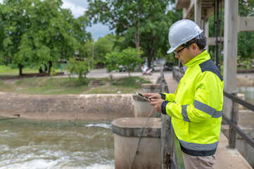 A engineering doing his checking routine. He is wearing hard hat and engineer uniform.Standing by the rail by the dam.Monitor water levels from the heavy rain that has been falling for several days.