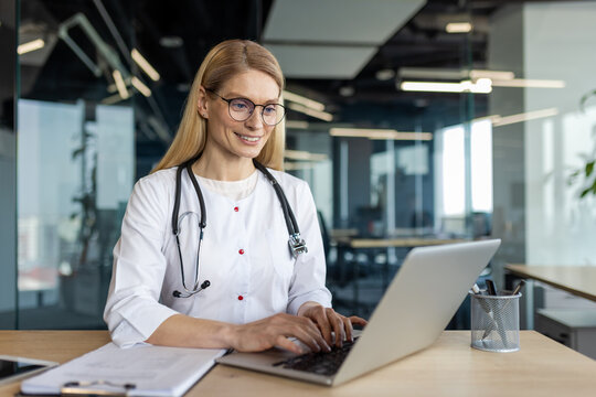 Confident female doctor working on laptop in office. Woman wearing glasses and stethoscope, engaging in online communication. Health professional using technology for medical consultations.